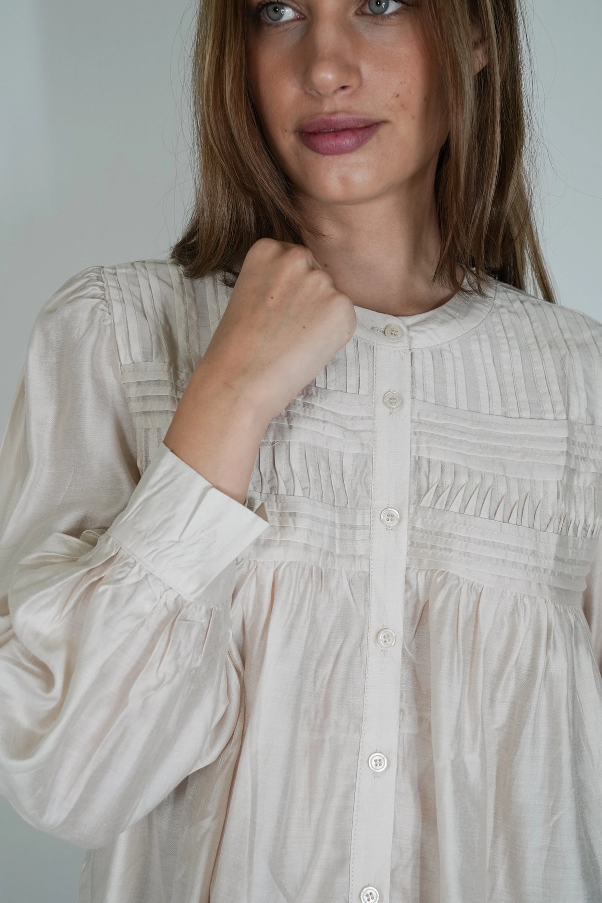 A close-up of a woman wearing a cream-colored, long-sleeved blouse with vertical pleating and a button-down front.