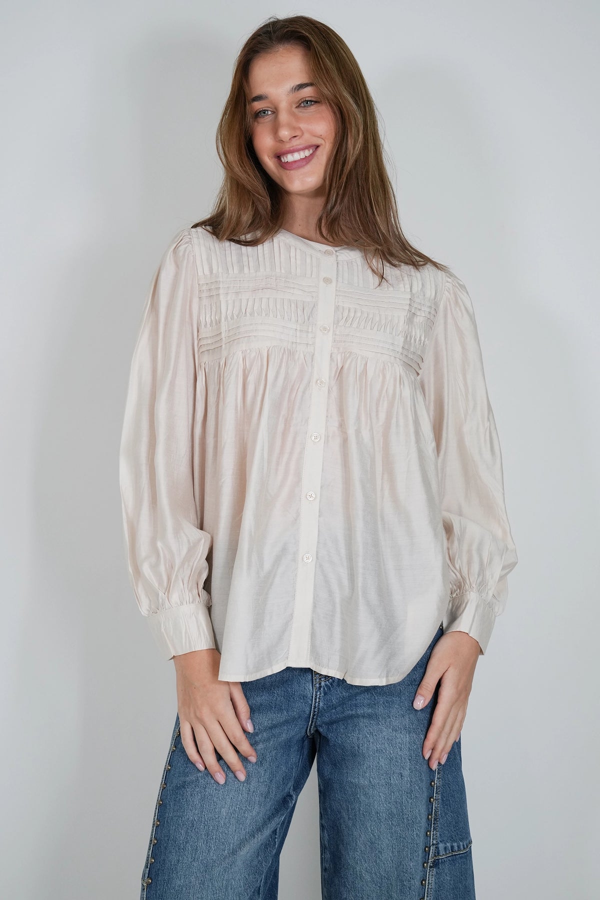 A woman wearing a cream-colored long-sleeve blouse with a gathered, smocked bodice and button-down front, paired with blue jeans, standing against a plain light gray background.