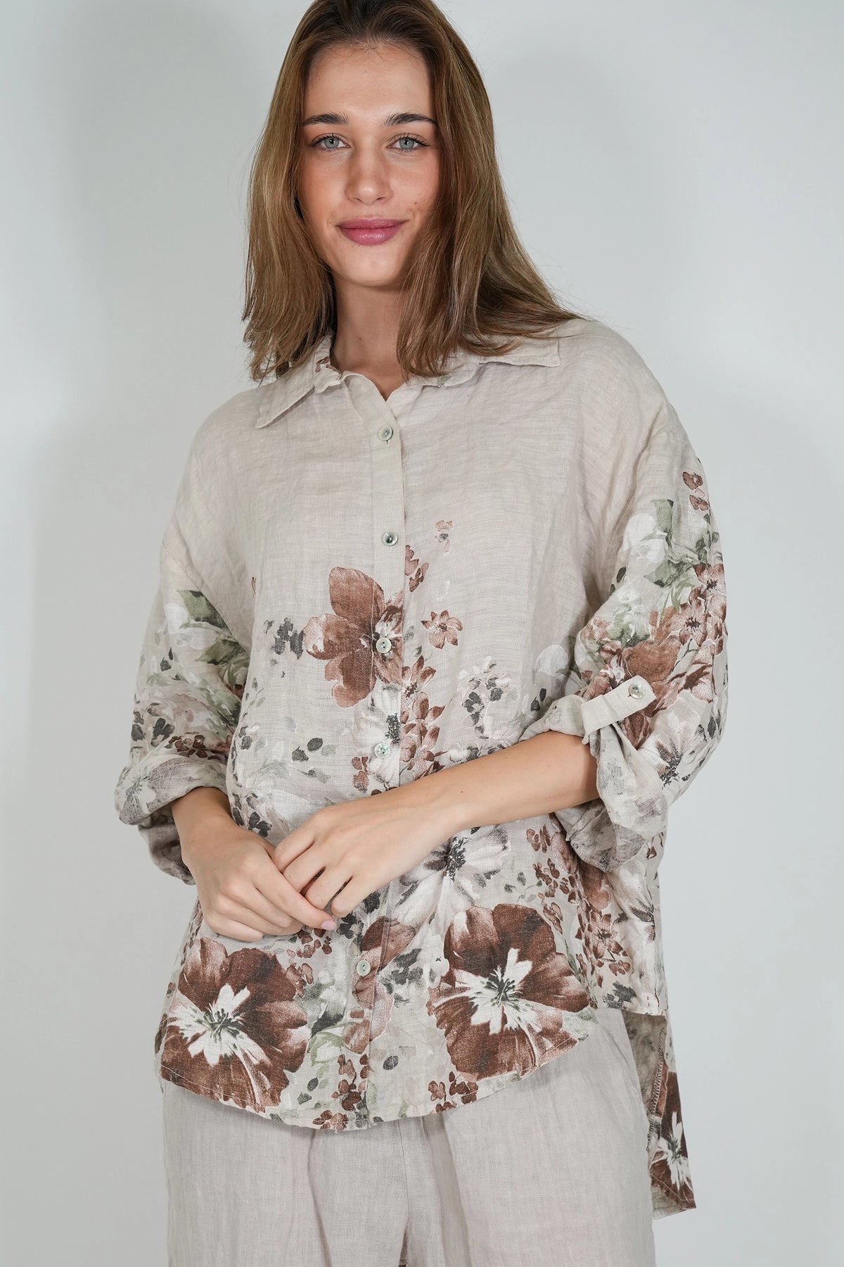 A woman wearing a light beige linen button-up blouse with a brown and white floral print, paired with light beige trousers, photographed against a neutral light gray background.
