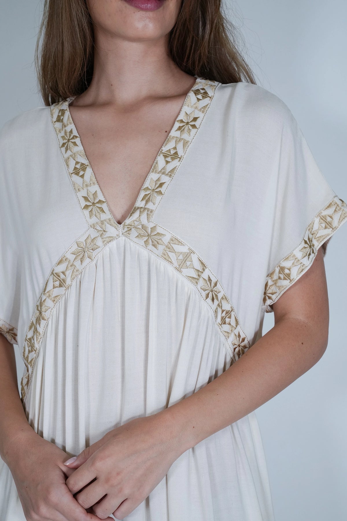 A close-up of a woman wearing a cream-colored kaftan dress with gold embroidered details along the V-neckline and sleeve cuffs, set against a plain light gray background.