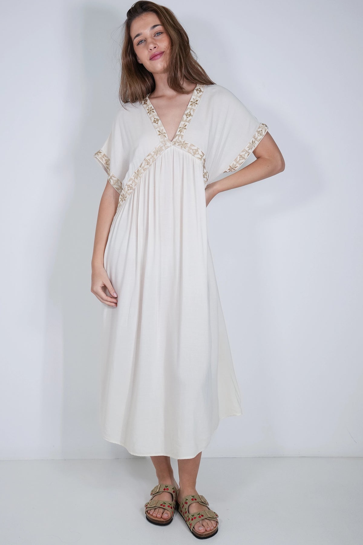 A woman modeling a flowing off-white maxi dress with short flutter sleeves and gold embroidered trim along the neckline and sleeve edges, paired with brown strappy sandals against a plain white studio background.