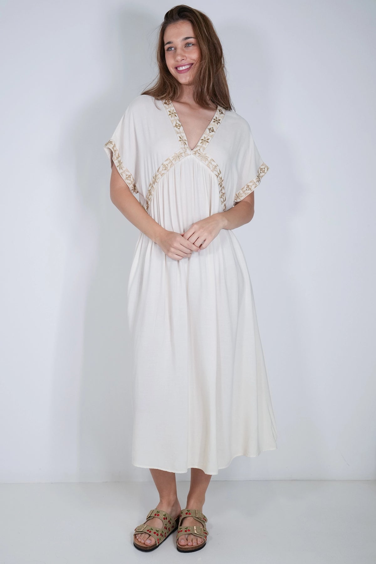 A woman modeling a loose, off-white midi dress with gold embroidered trim along the neckline and sleeve cuffs, paired with brown strappy sandals, set against a plain white studio background.
