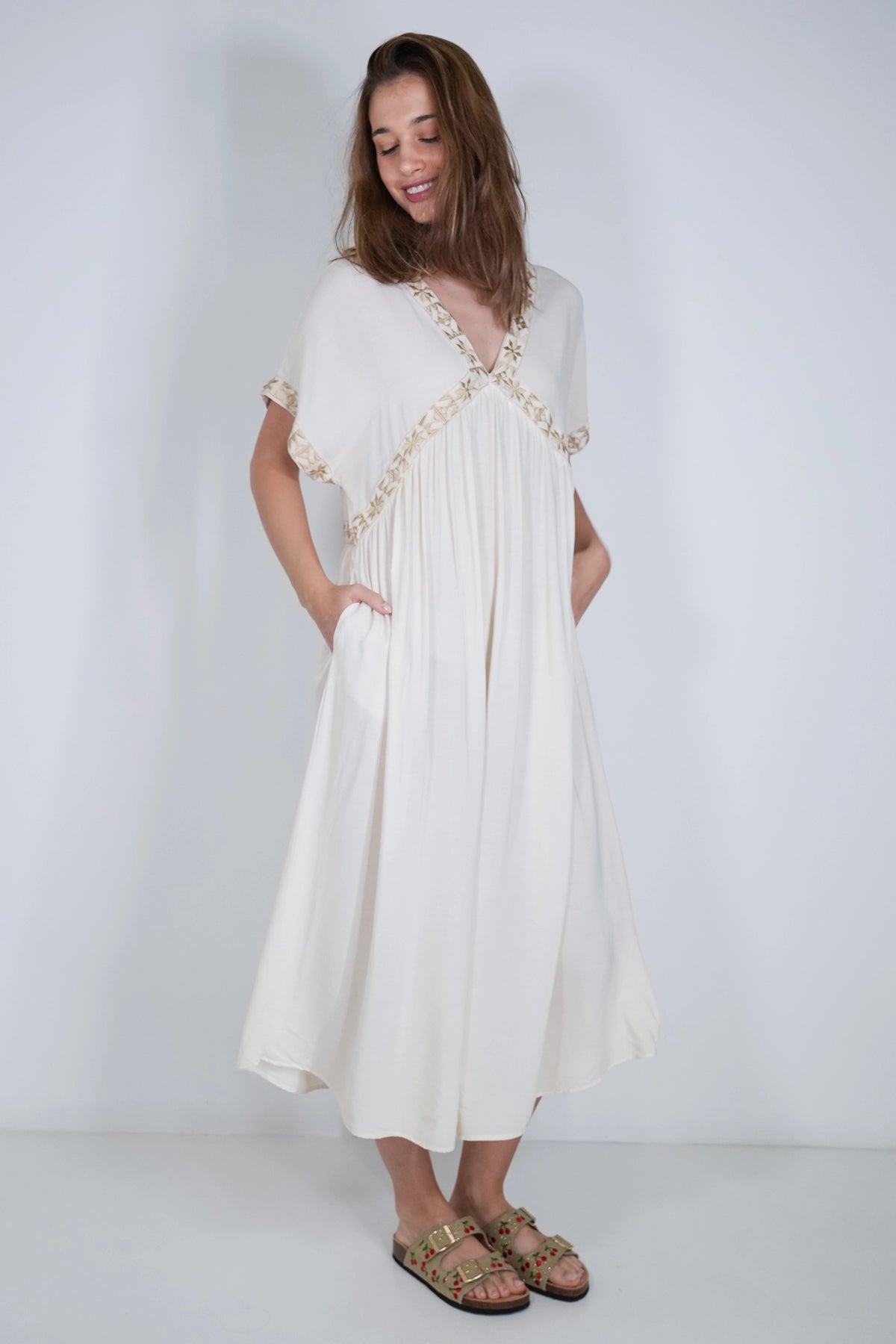 A woman modeling a cream-colored midi dress with gold embroidered details along the V-neckline and sleeve cuffs, paired with brown sandals, set against a plain light gray studio background.