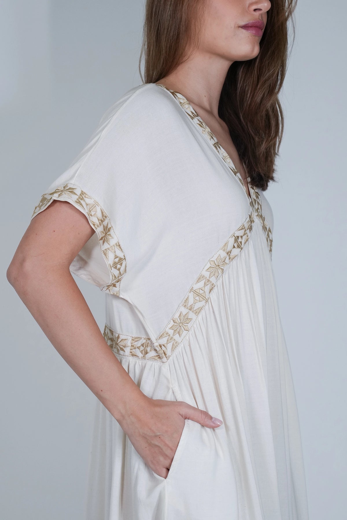 A woman wearing a loose white or off-white dress with gold embroidered trim along the neckline, sleeve edges, and waist, photographed against a plain light gray background.