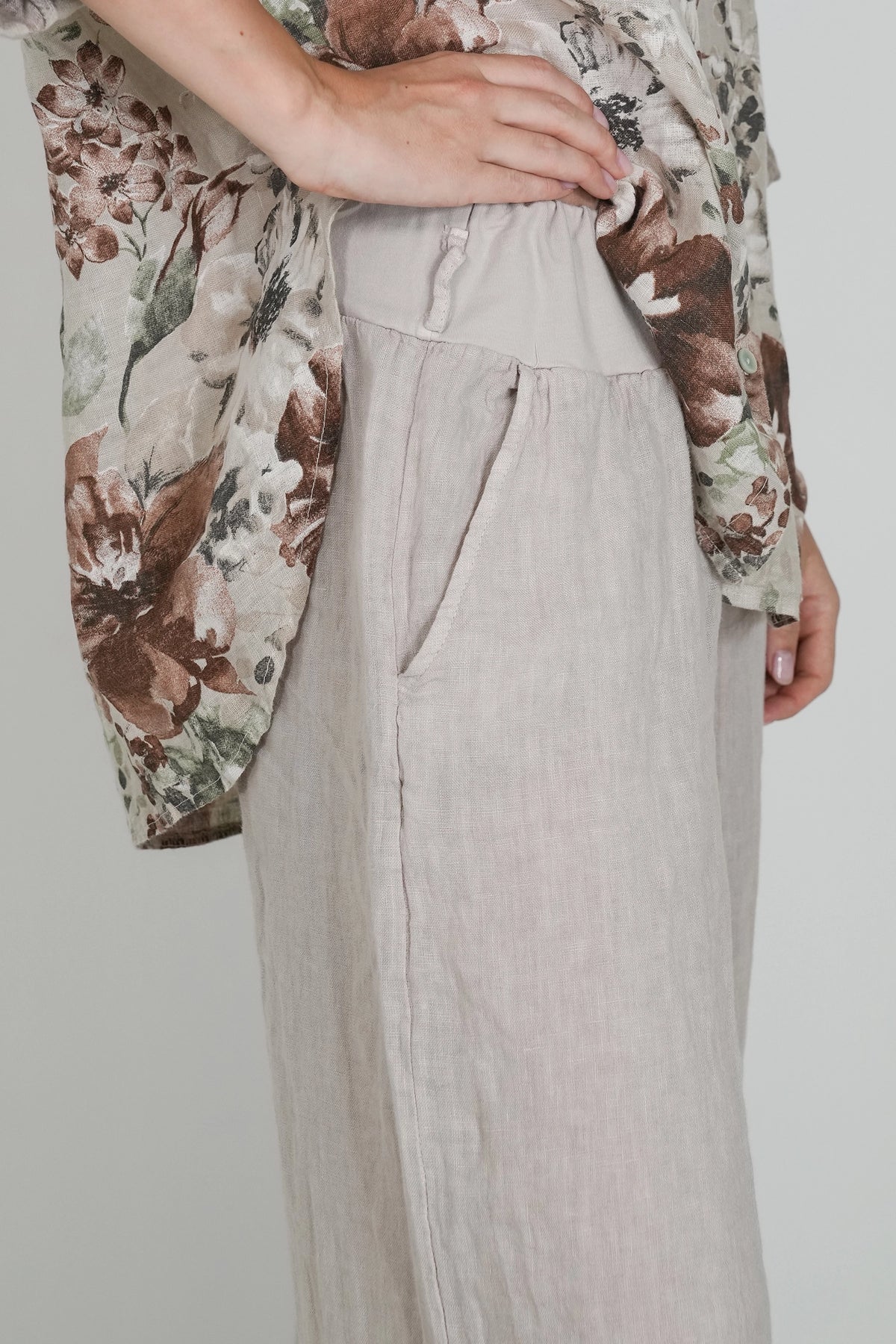 A close-up of a person wearing a light beige linen skirt with side pockets and a matching waistband, paired with a floral printed top. The background is a solid light gray.