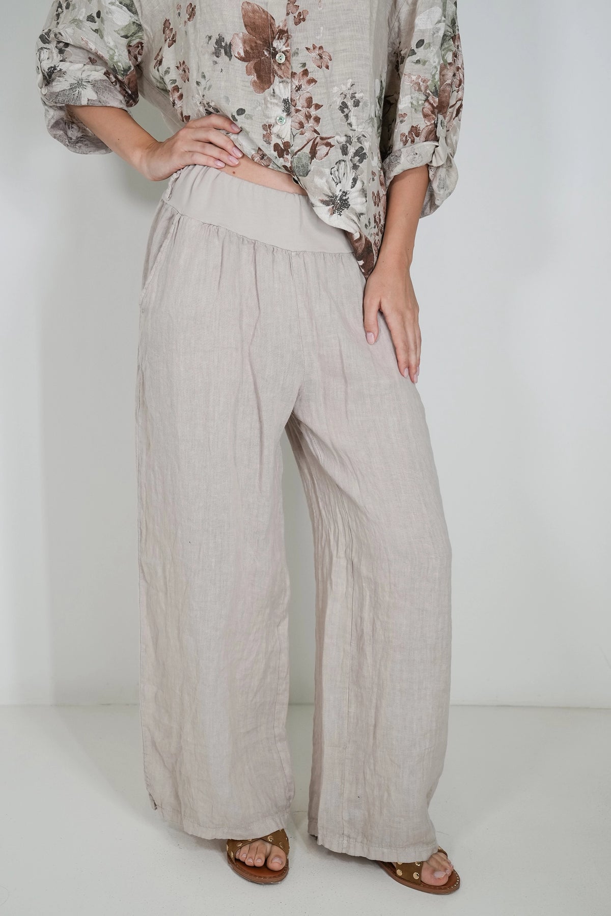 A person wearing loose, wide-leg linen pants in a light beige color, paired with a floral long-sleeve top and brown sandals, standing against a plain white studio background.