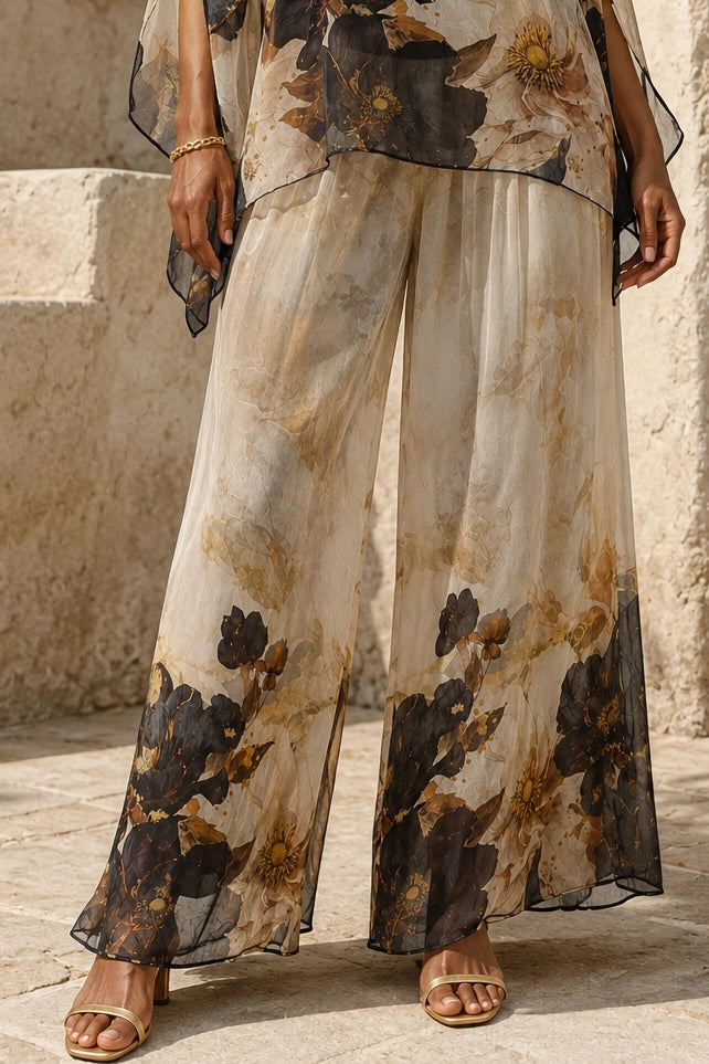 A person wearing sheer wide-leg pants with a beige, brown, and black floral print, paired with a matching sheer top and gold-toned sandals, standing outdoors against a stone wall.