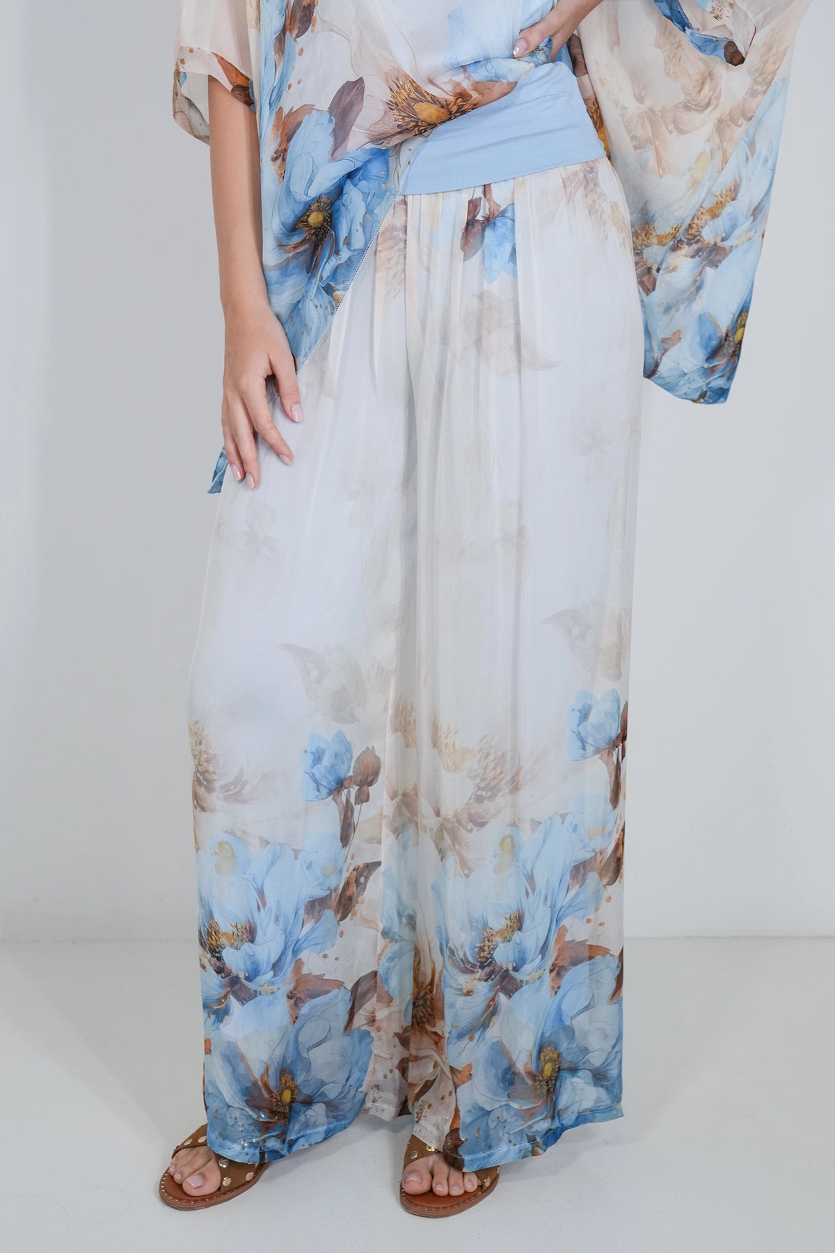 A model wearing a white wide-leg maxi dress with a blue and brown floral print, styled with a light blue sash and brown sandals.