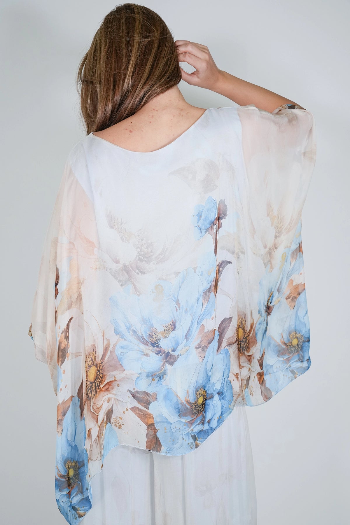 A woman shown from behind wearing a sheer white kaftan-style top with a large blue and beige floral print, set against a plain light gray background.