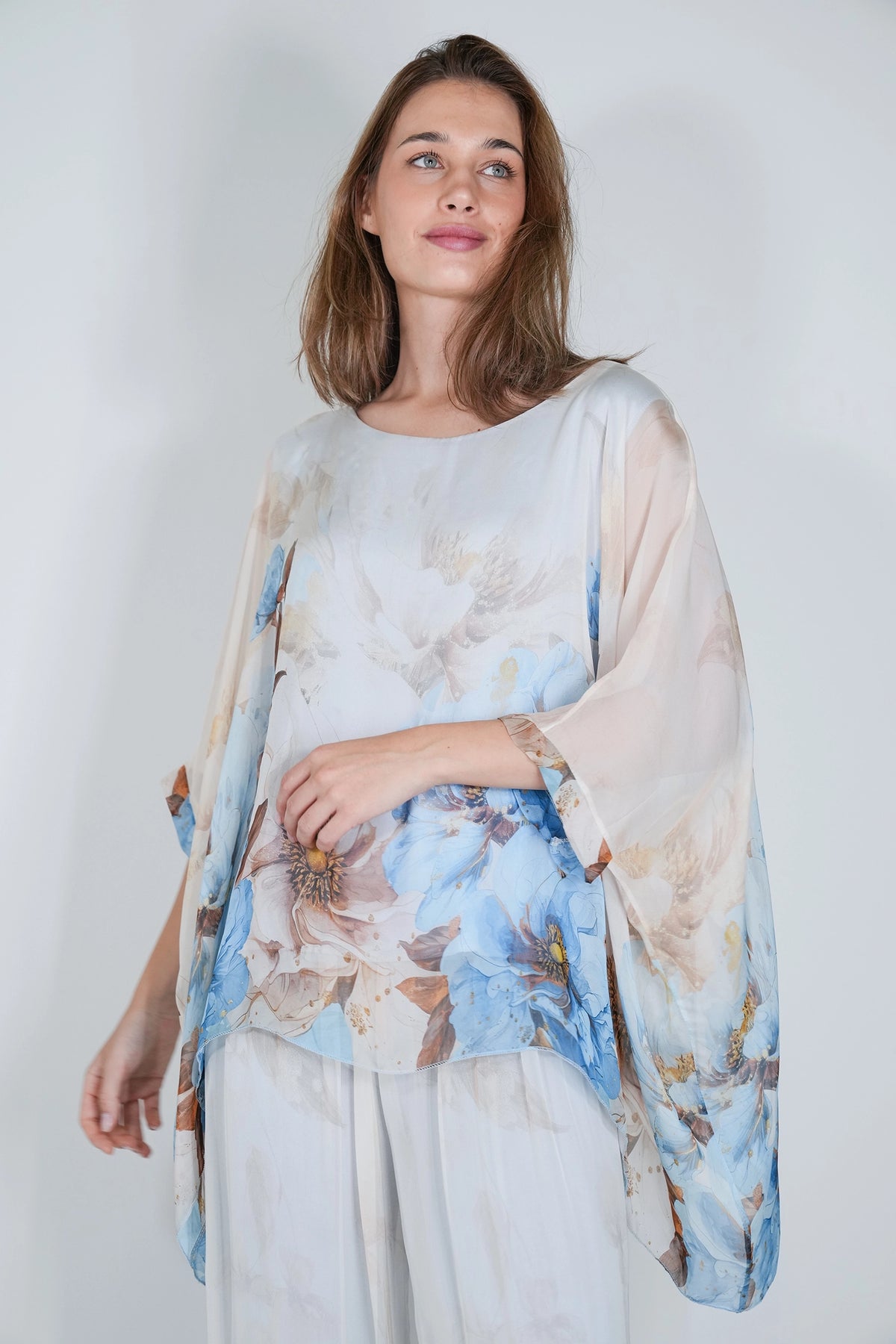 A woman wearing a sheer, lightweight floral top with wide draped sleeves and light-colored trousers, photographed against a plain light gray studio background.