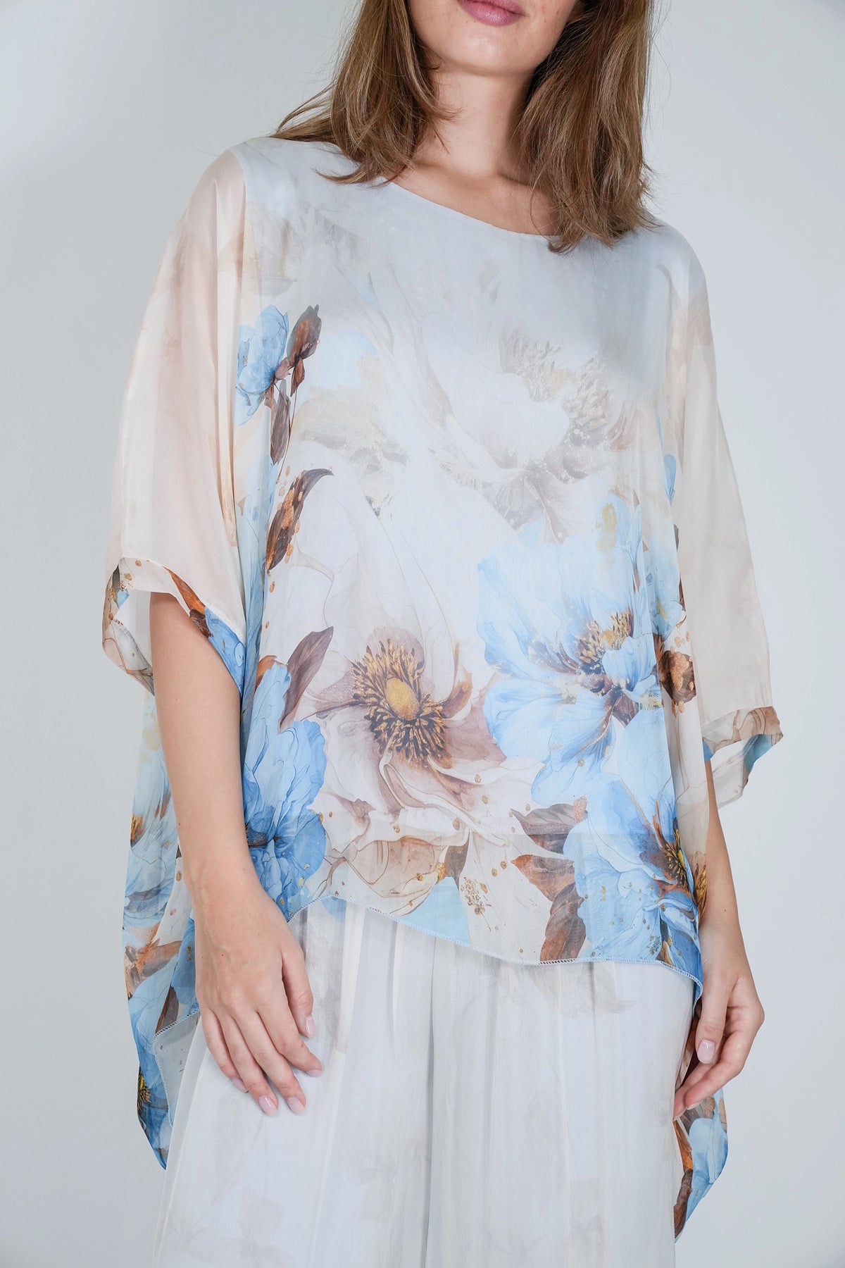 A woman wearing a loose, semi-sheer floral top with wide short sleeves and a round neckline, paired with white pants, against a plain light gray background.