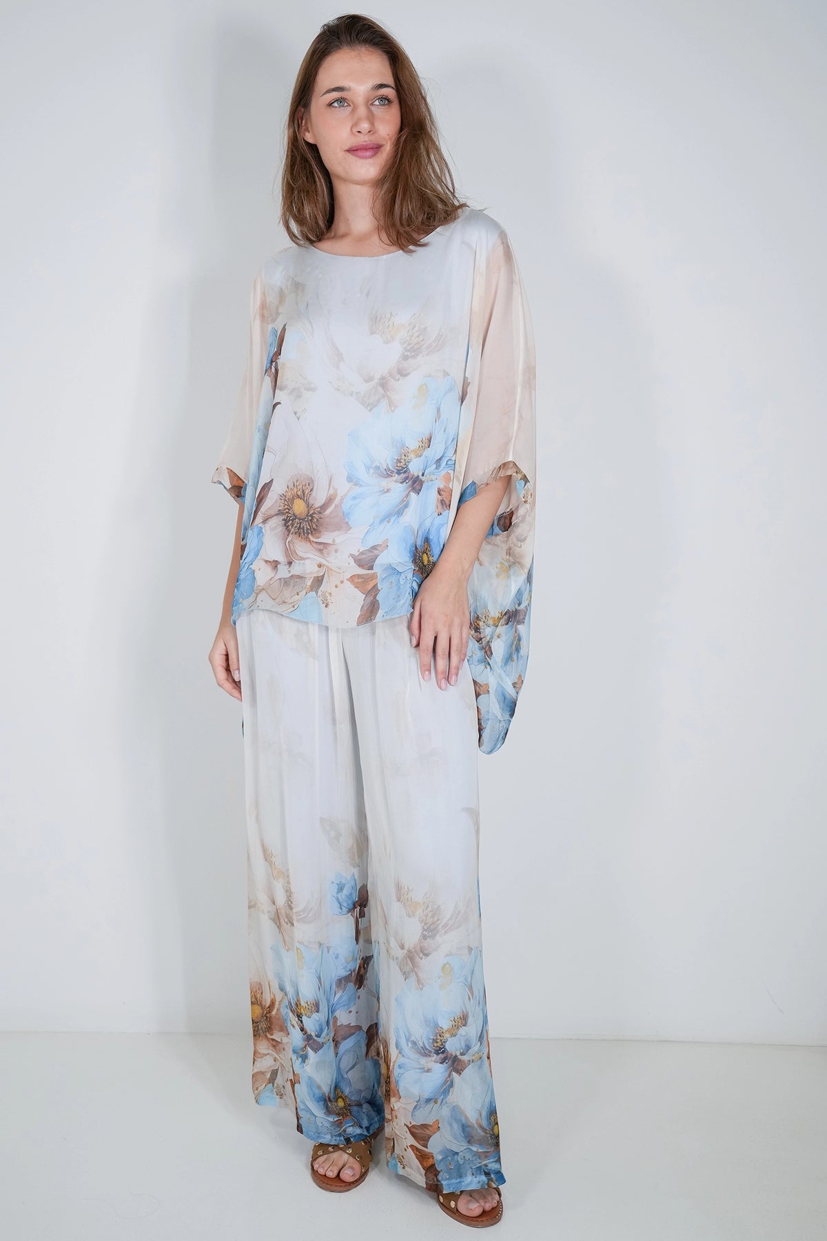 A woman modeling a matching two-piece sheer floral set consisting of a long-sleeve top and wide-leg pants, in light blue, beige, and brown tones, set against a plain white studio background.