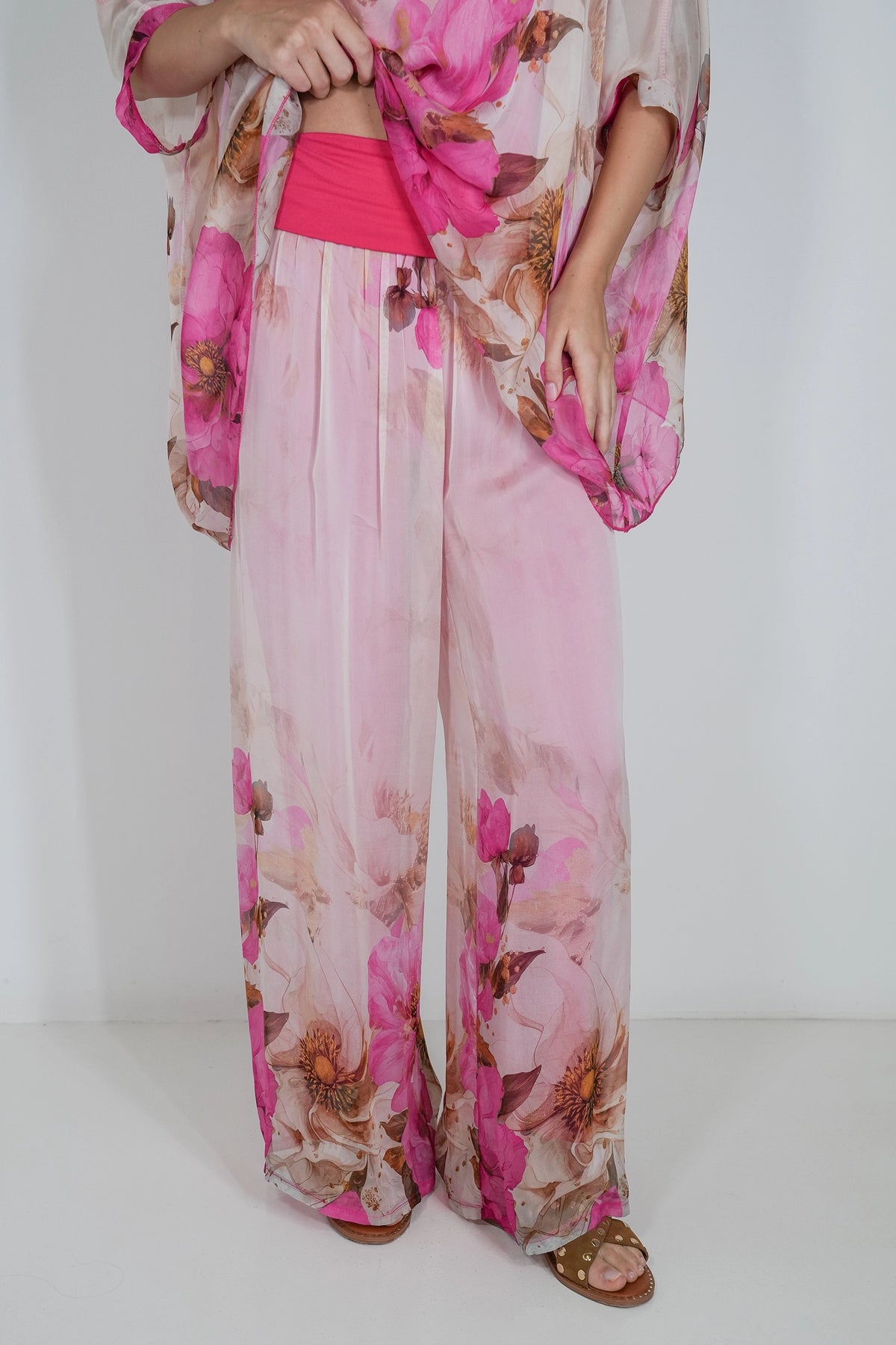 A person wearing wide-leg pink floral pants with a sheer pink top and matching outer layer, standing against a plain light background.