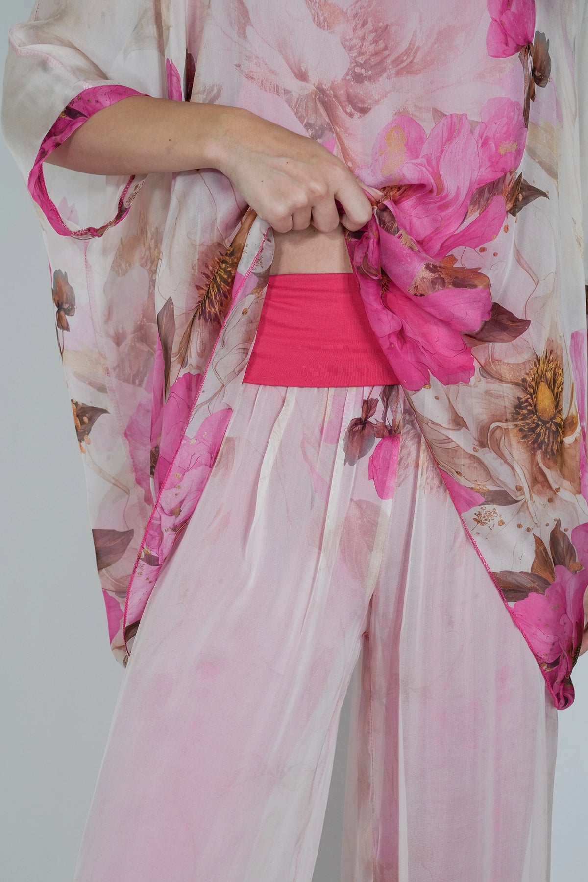 A close-up of a person wearing a sheer light pink floral kimono-style top with large pink and mauve flowers, paired with a bright pink waistband or undergarment, against a plain light gray background.