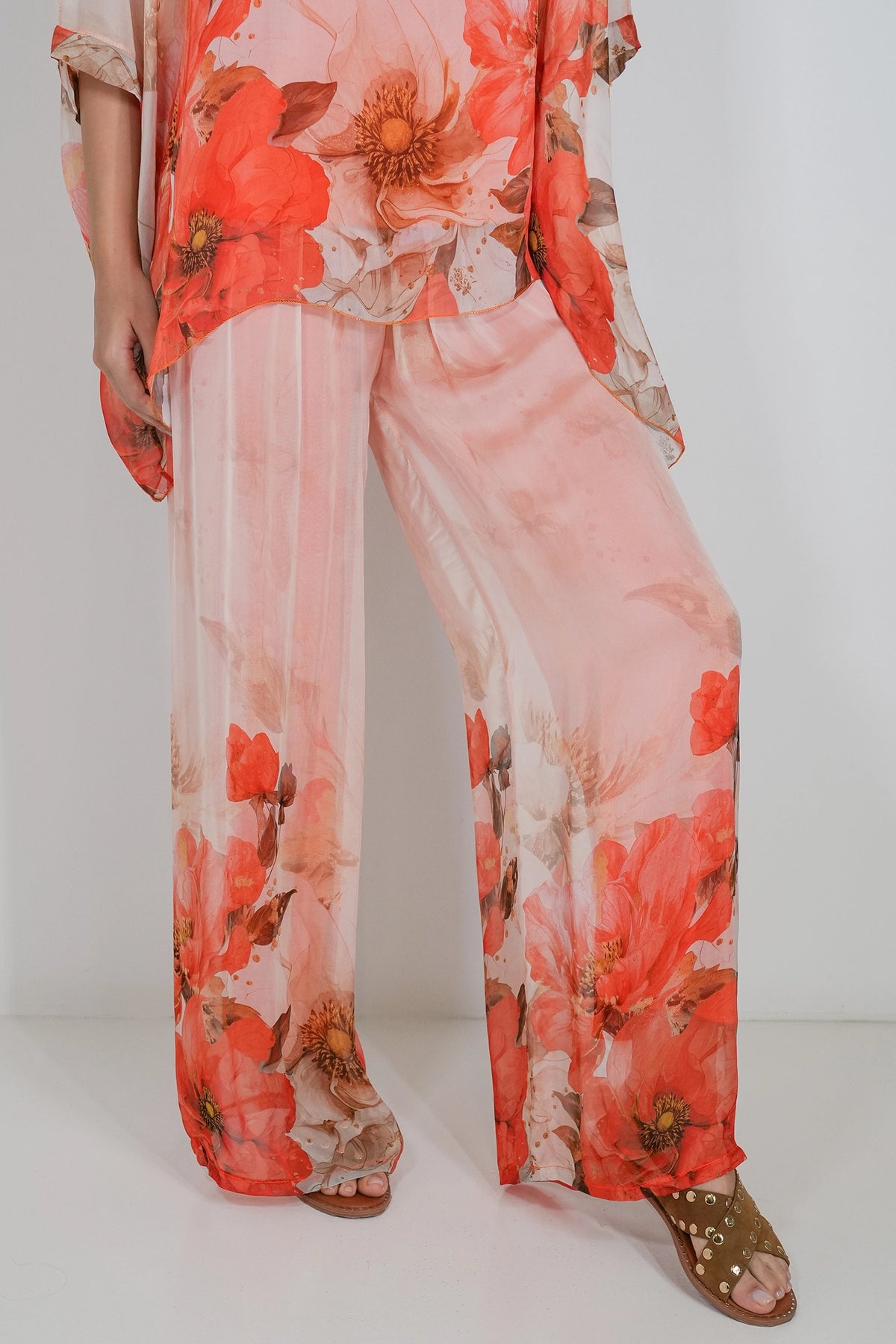 A model wearing a sheer, light pink two-piece set with a vibrant red and orange floral print, paired with brown studded sandals, shown from the waist down against a plain white background.