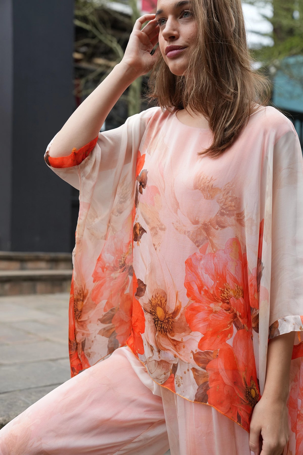 A woman wearing a loose, semi-sheer peach and pink kaftan-style top with large orange and red floral prints, paired with matching pink pants, photographed outdoors.
