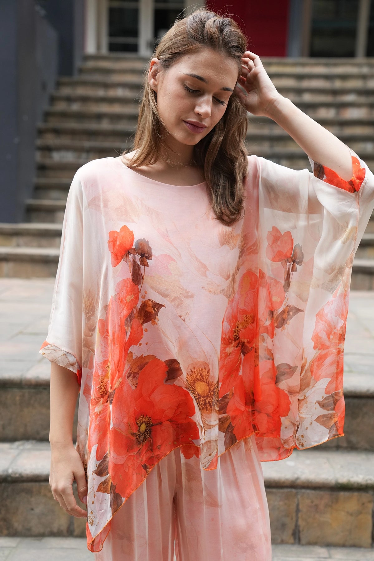A woman wearing a sheer, light pink kaftan-style top with a large red and orange floral print, paired with matching pink trousers, standing on stone steps.