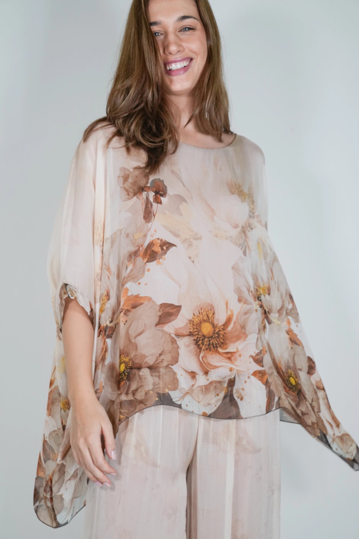 A woman wearing a sheer, lightweight beige top with a large brown and tan floral print, paired with matching sheer pants, set against a plain light gray background.