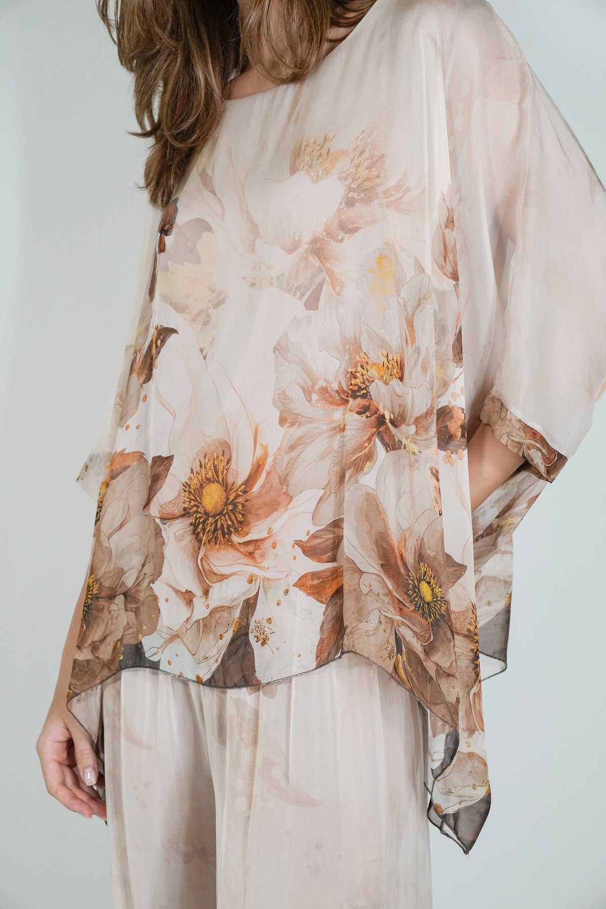 A woman wearing a sheer, lightweight top with a large floral print in soft beige, brown, and muted orange tones, paired with light beige pants against a plain light background.