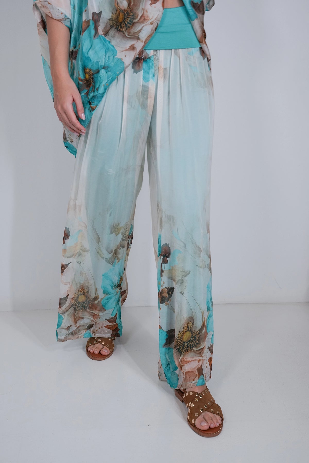 A person wearing wide-leg sheer pants with a turquoise, beige, and brown floral print, paired with a matching top and brown sandals, photographed against a plain light gray studio background.