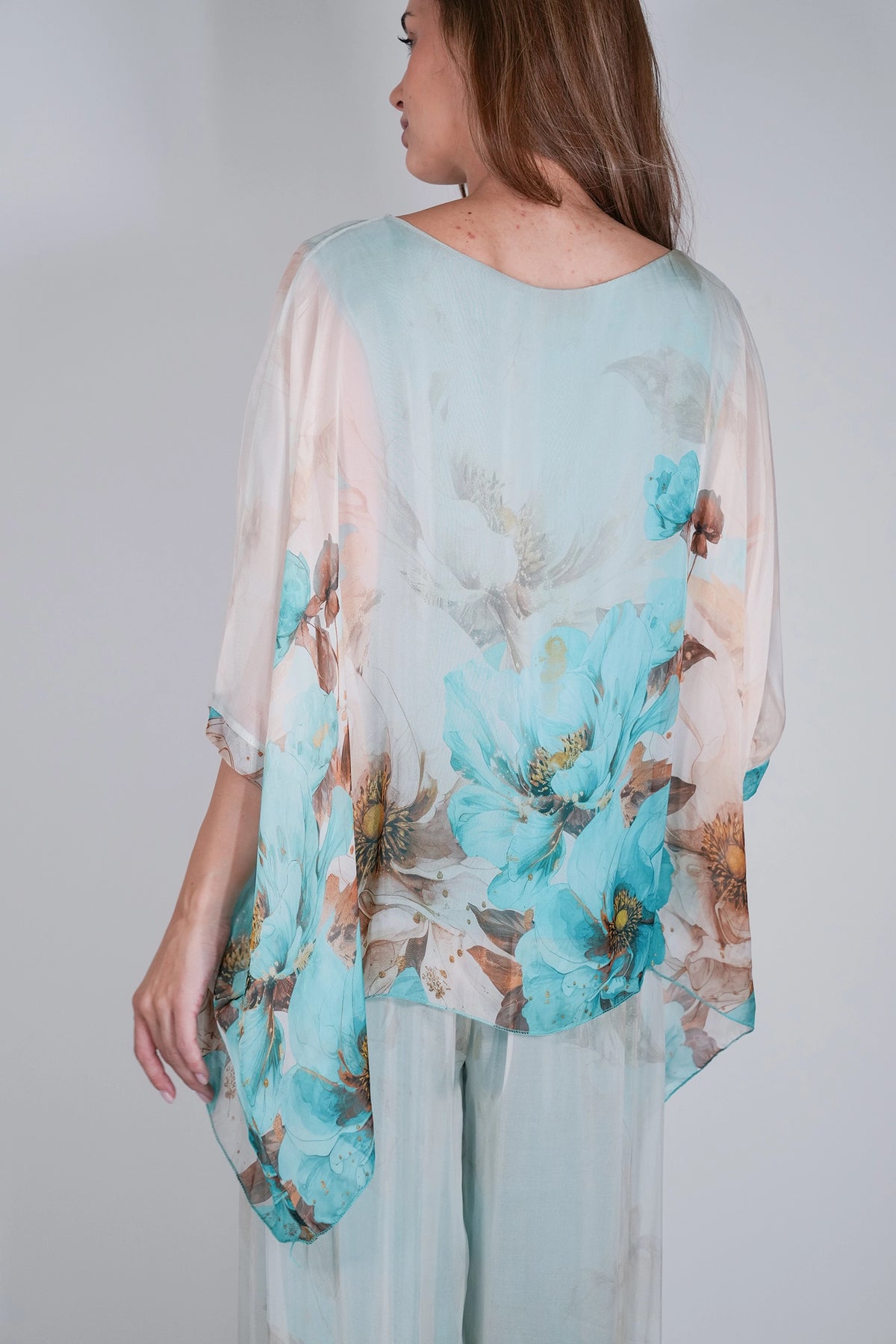 A woman shown from the back wearing a sheer, flowing top with a large floral print in turquoise, beige, and brown over a pale mint base, paired with matching sheer pants against a light gray studio background.