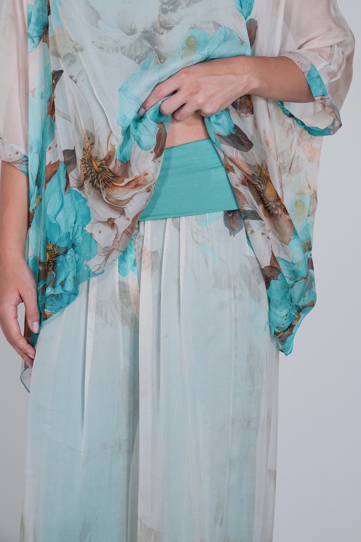 A close-up of a model wearing a light blue and white floral print kaftan-style tunic with a solid turquoise waistband and matching turquoise sash, paired with light blue wide-leg trousers.