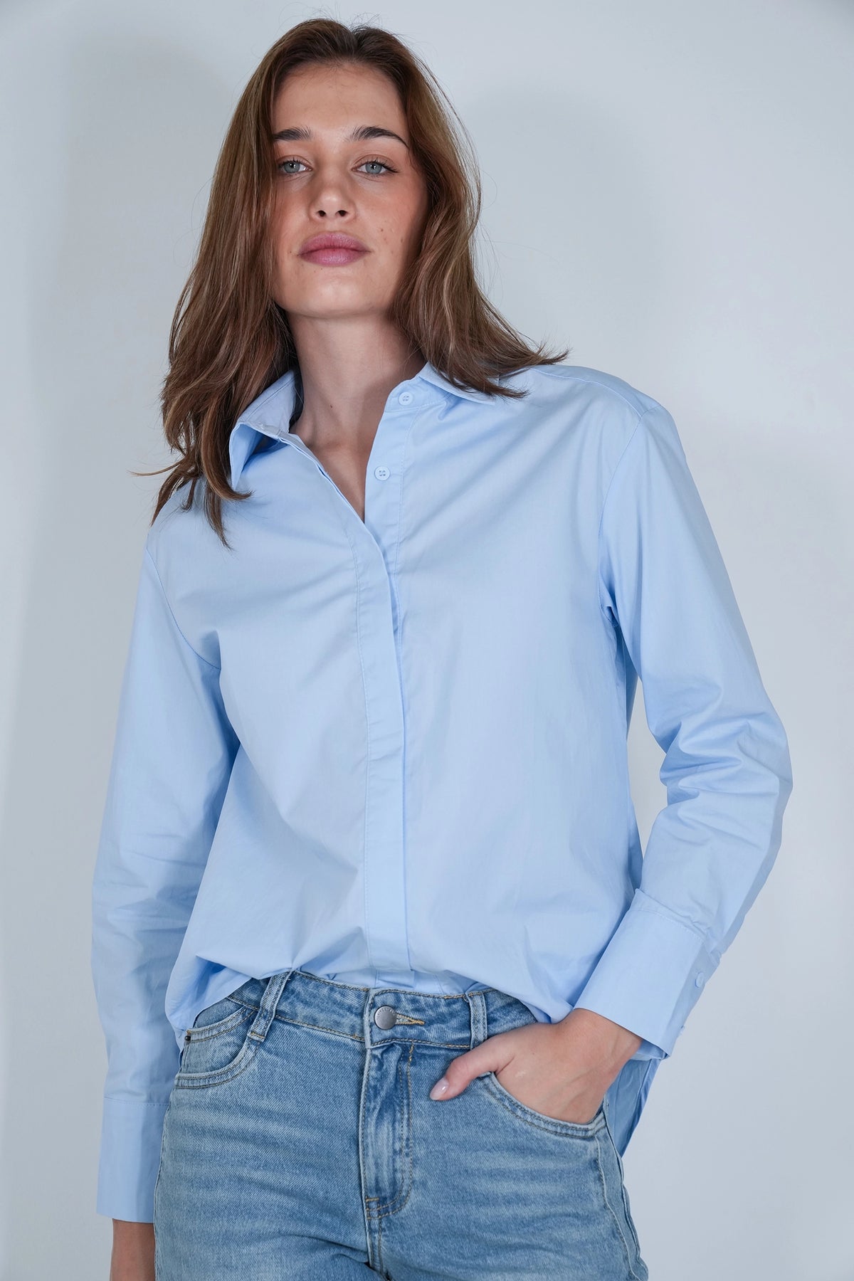 A woman wearing a light blue long-sleeve button-up shirt and light-wash blue jeans, standing against a plain light gray studio background.