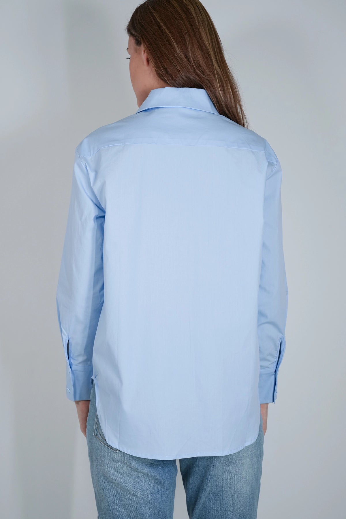 A woman viewed from the back wearing a light blue long-sleeve button-up shirt and blue jeans against a plain light gray studio background.