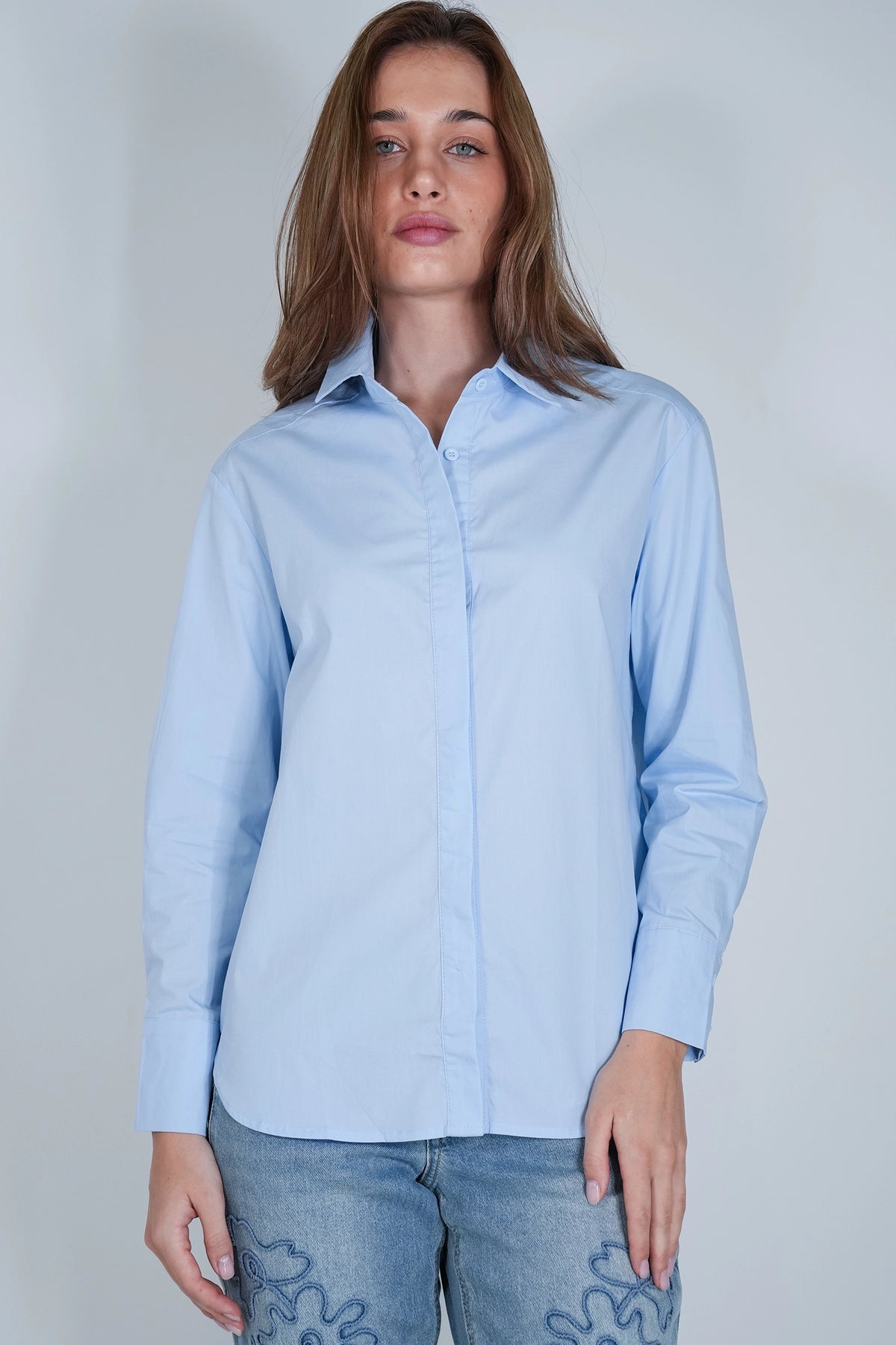 A woman wearing a light blue long-sleeve button-up shirt and light-wash jeans, standing against a plain light gray studio background.