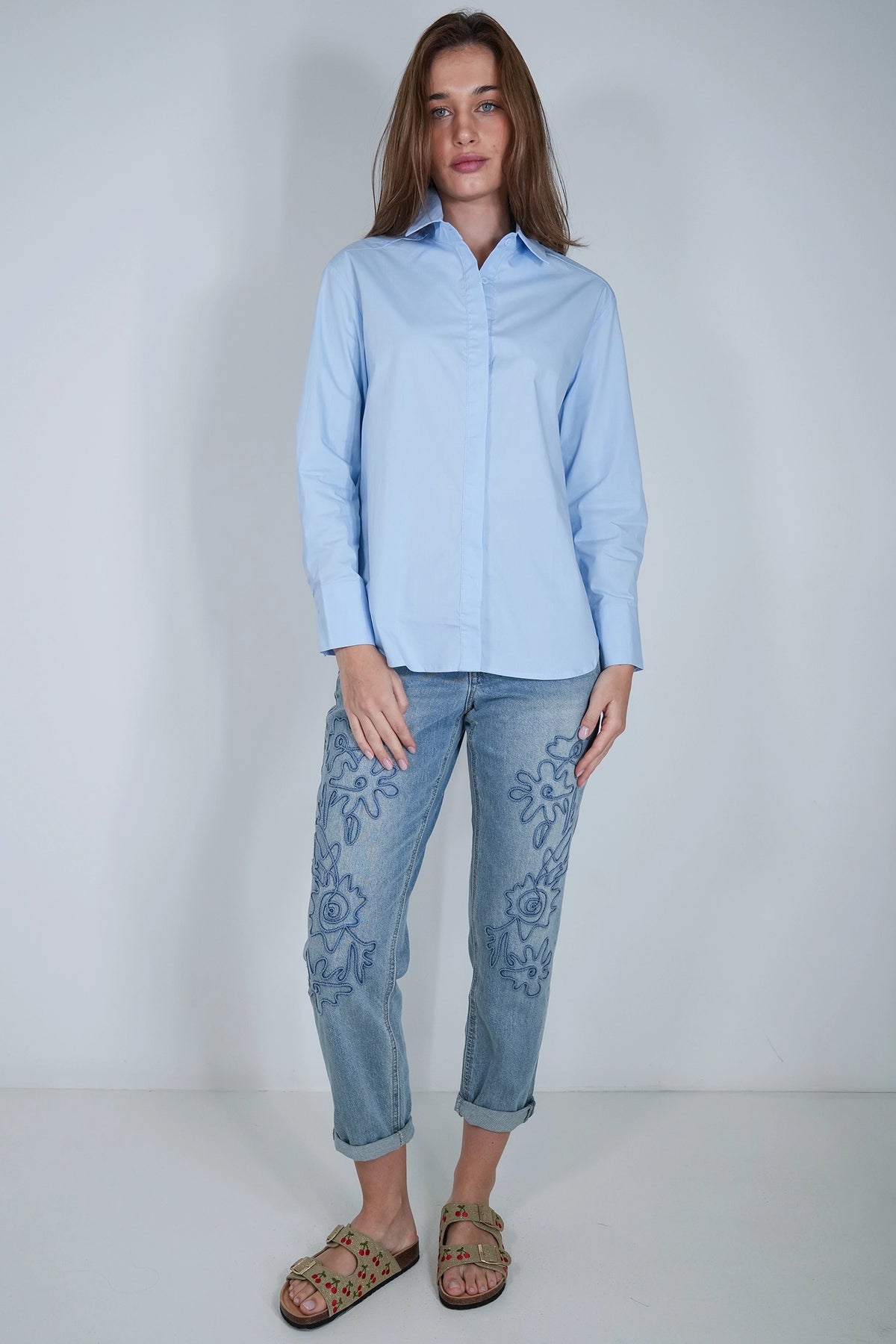 A woman modeling a light blue long-sleeve button-up shirt and light-wash blue jeans with floral embroidery on the legs, paired with brown sandals, against a plain light gray studio background.