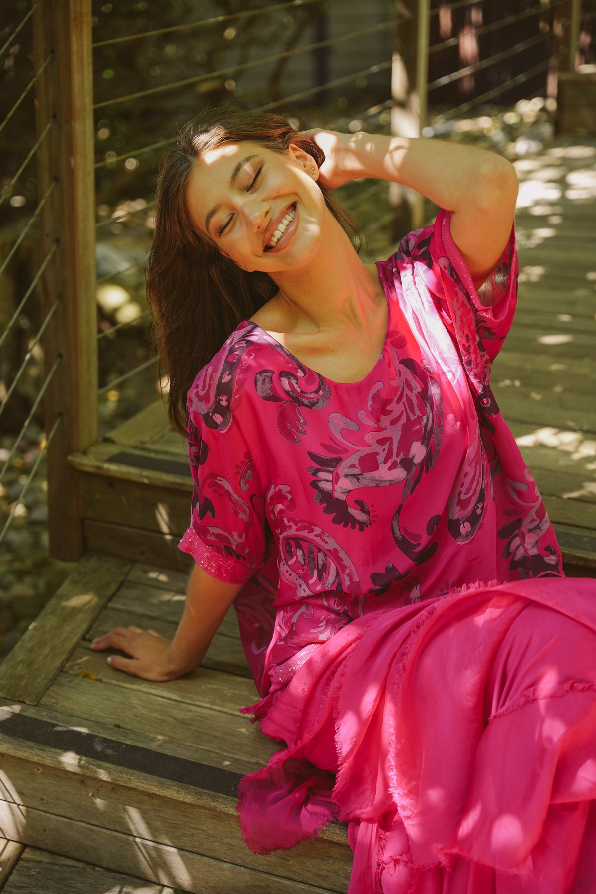 Woman in a pink dress with black floral pattern sitting on a wooden bench.