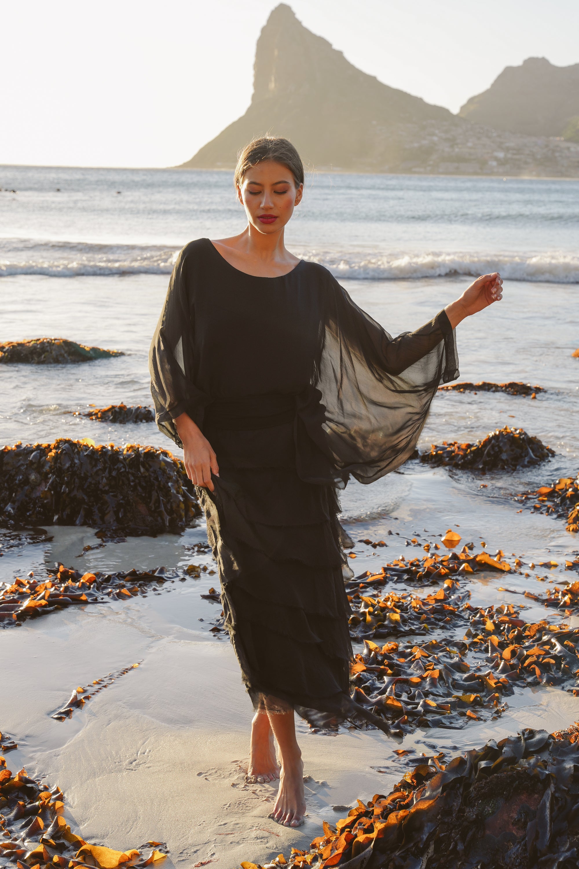 Woman in a black dress standing on a beach with mountains in the background