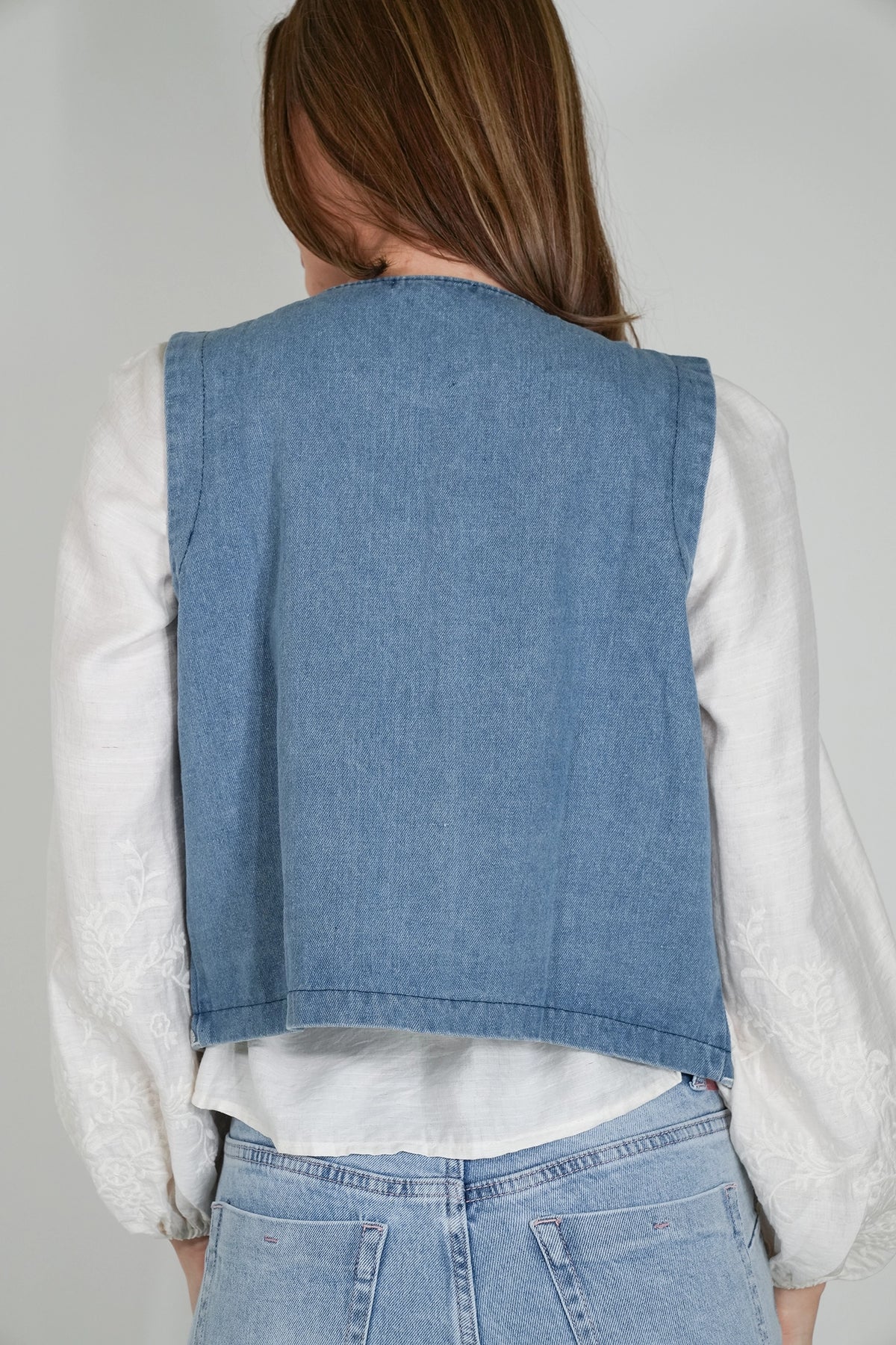 A woman shown from the back wearing a sleeveless light blue denim vest over a white long-sleeve blouse and light blue jeans, against a plain light gray studio background.