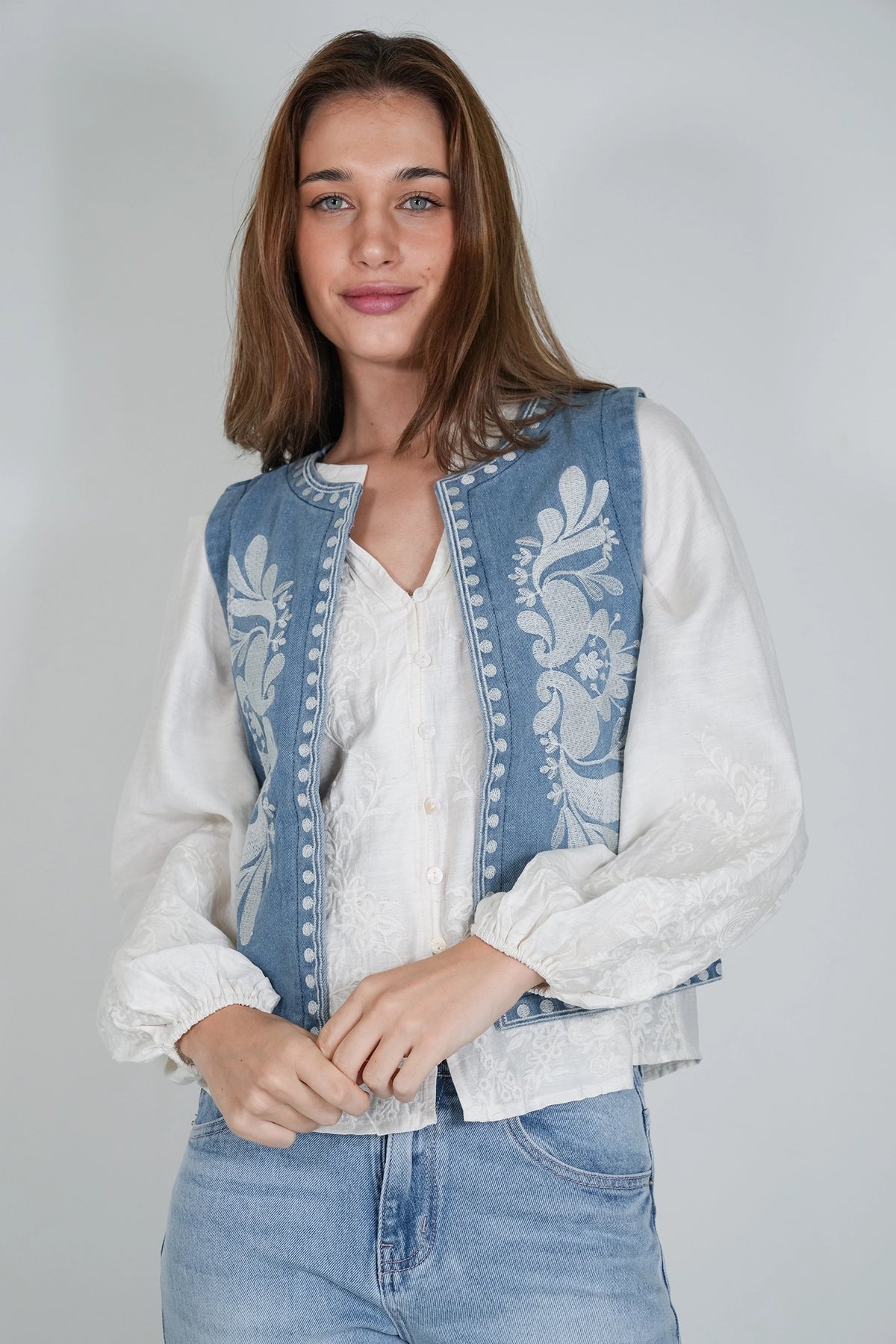 A woman wearing a light blue denim vest with white floral embroidery over a white lace blouse and light blue jeans, photographed against a plain light gray studio background.