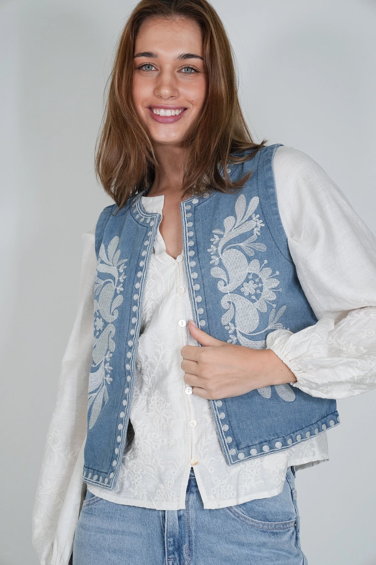 A smiling woman wearing a light blue denim vest with white floral embroidery and silver-tone studs, layered over a white lace blouse and paired with light blue jeans, against a light gray studio background.