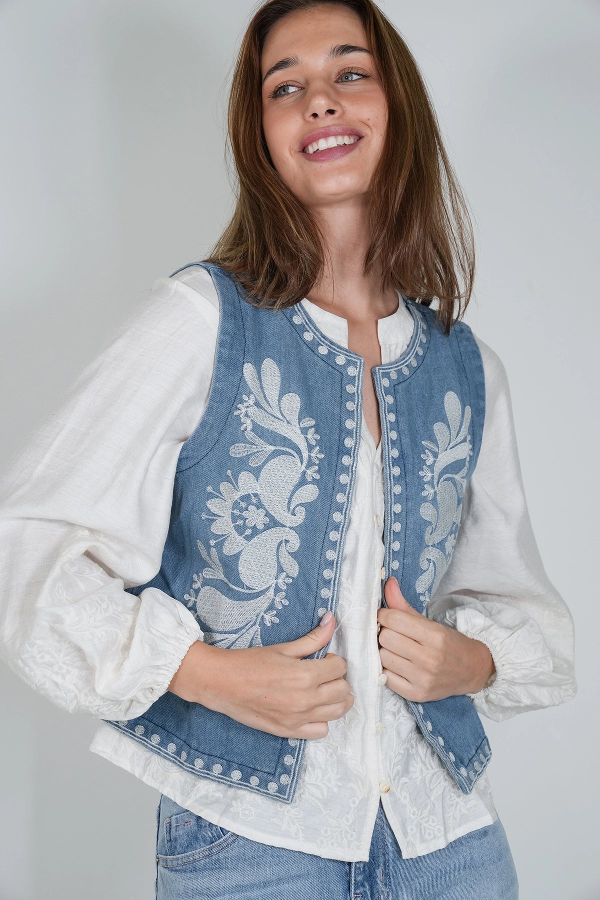 A woman wearing a light blue denim vest with white floral embroidery over a white long-sleeve blouse and blue jeans, photographed against a plain light gray studio background.