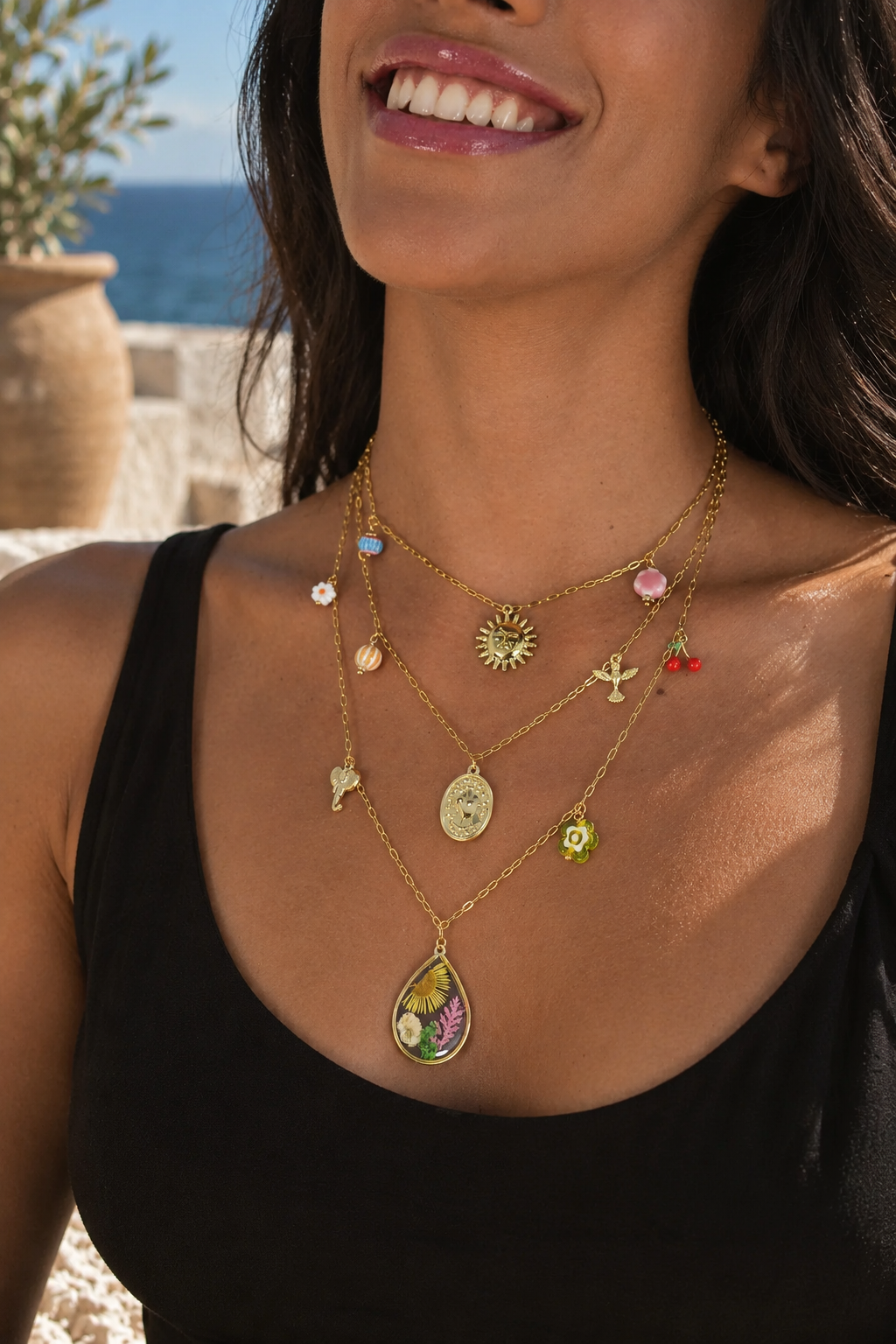 A close-up lifestyle shot of a woman wearing a layered gold necklace set with various colorful charms, including a teardrop pendant filled with dried flowers, against a sunny outdoor background.