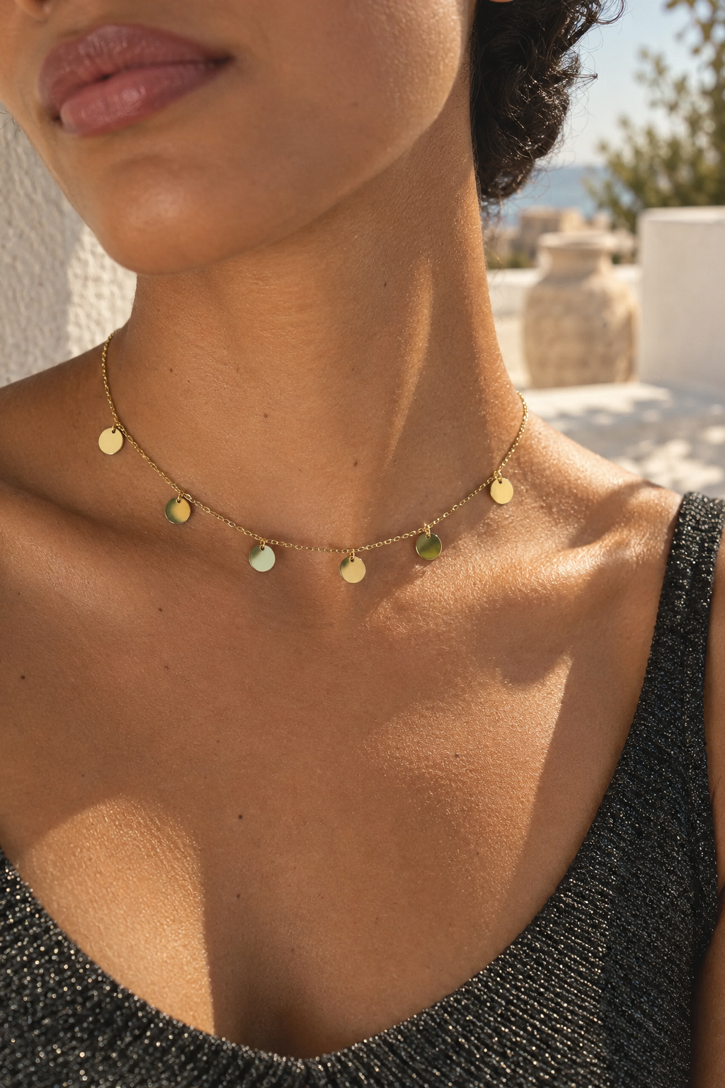 A close-up lifestyle shot of a model wearing a delicate gold chain necklace with small, irregularly shaped pastel-colored gemstone pendants in green, blue, and white, set against a warm outdoor background.