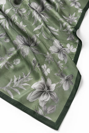 A close-up of a green silk scarf with a gray and white floral print and a solid dark green border, laid out on a white background.