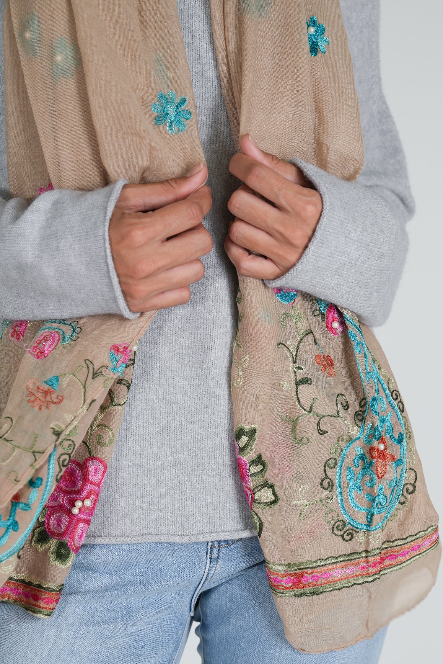 Person wearing a beige scarf with floral embroidery, gray sweater, and blue jeans on a white background