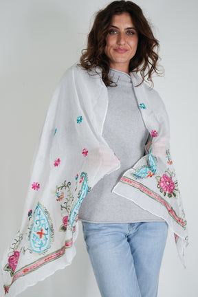 Woman wearing a white embroidered shawl over a gray sweater and light blue jeans on a plain background