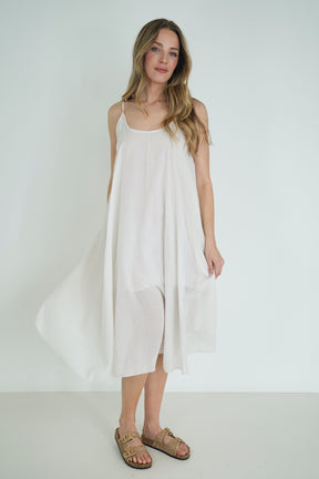 Model wearing a loose white strappy midi dress with flowy fabric, styled with gold sandals, posing against a plain white background.