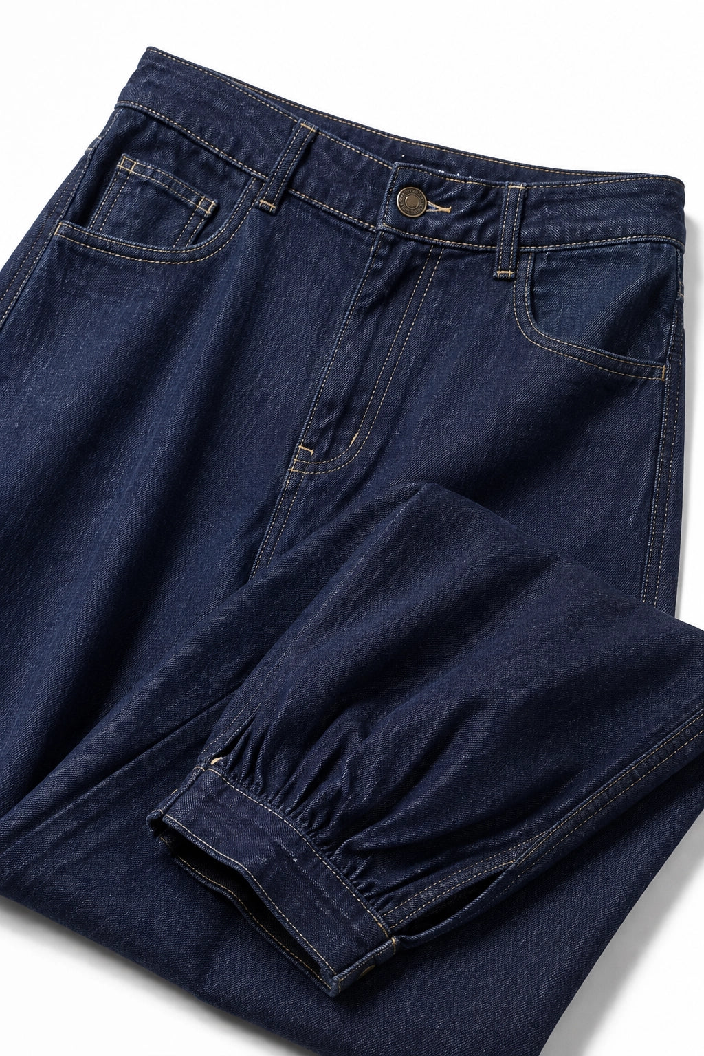 Dark indigo blue denim jeans folded on a white background, showing the front pocket, waistband, and gathered cuff detail.