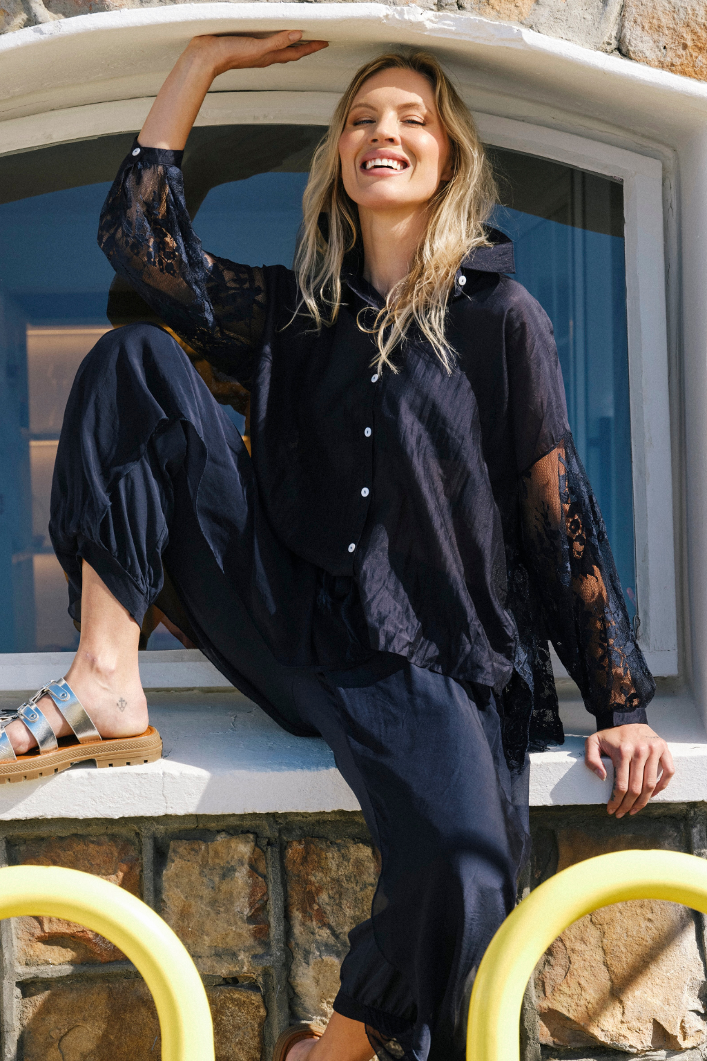 A woman models a black hooded button-up shirt with sheer lace sleeves and matching black pants, sitting on a stone ledge next to a window.