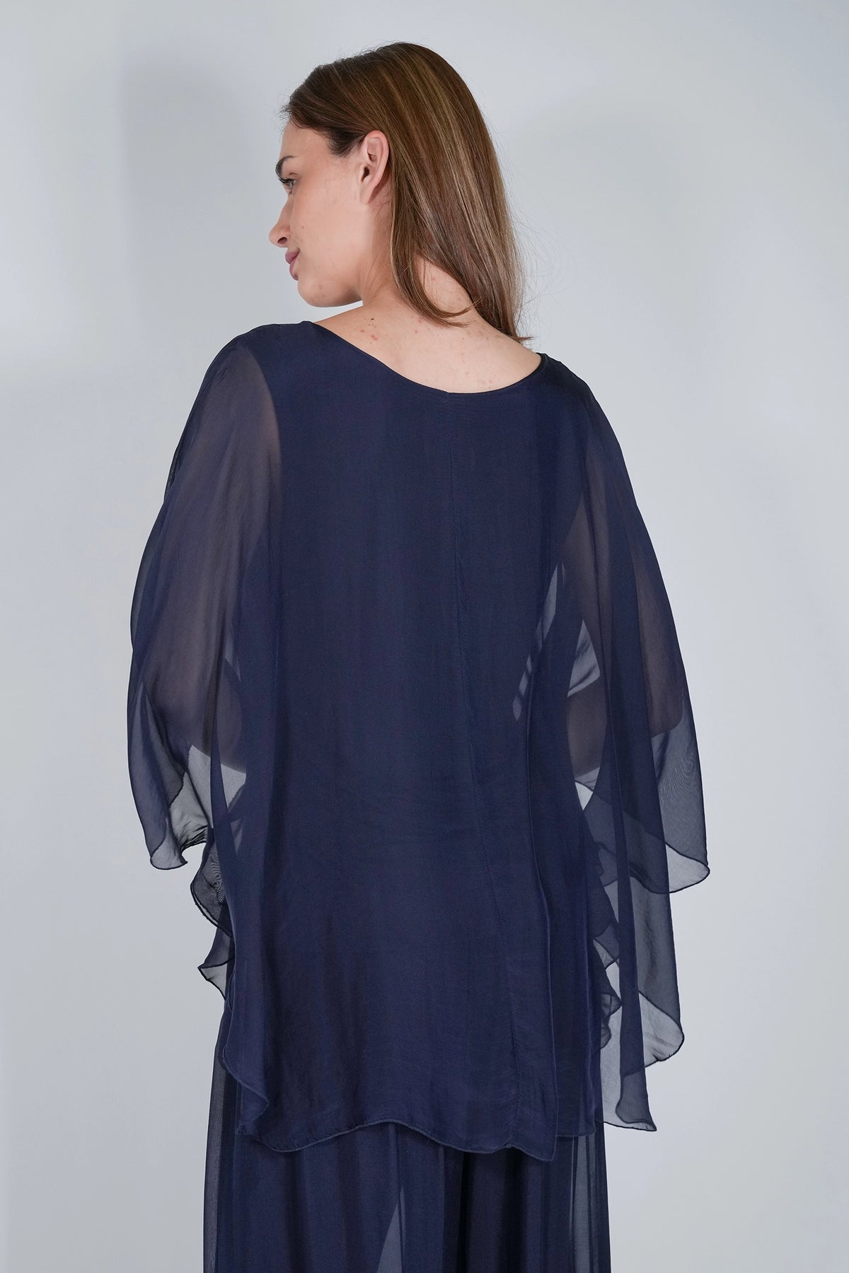 A woman shown from the back wearing a navy blue sheer draped top with wide flutter sleeves and a matching navy skirt, photographed against a plain light gray studio background.