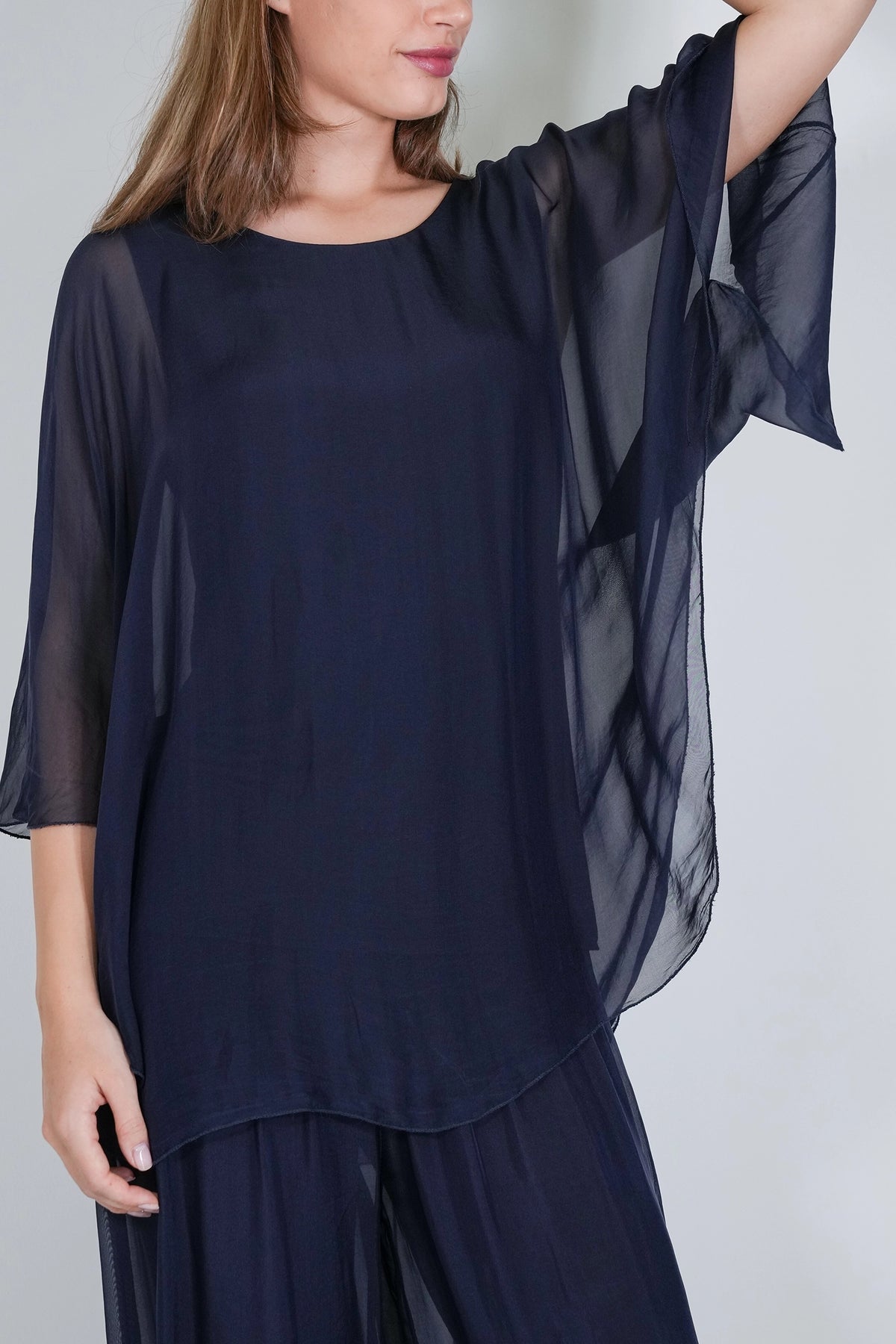 A woman wearing a navy blue sheer tunic top with wide three-quarter sleeves and matching navy pants, photographed against a light gray studio background.