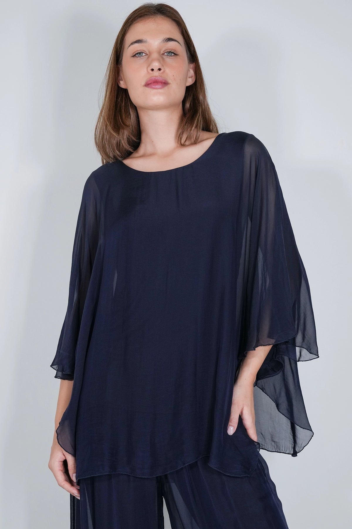 A woman wearing a loose, semi-sheer navy blue top with wide draped sleeves and matching navy pants, photographed against a light gray studio background.