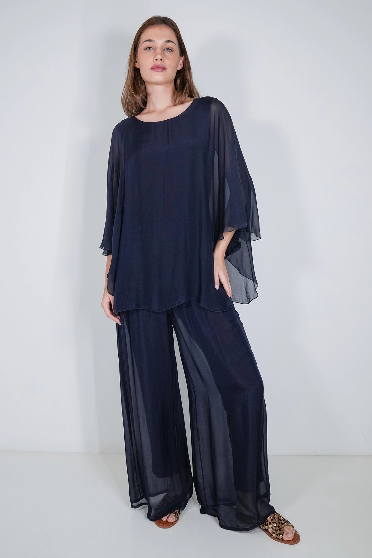 A woman modeling a matching navy blue sheer two-piece outfit consisting of a loose long-sleeve top with draped bell sleeves and wide-leg pants, paired with brown strappy sandals against a plain light gray studio background.