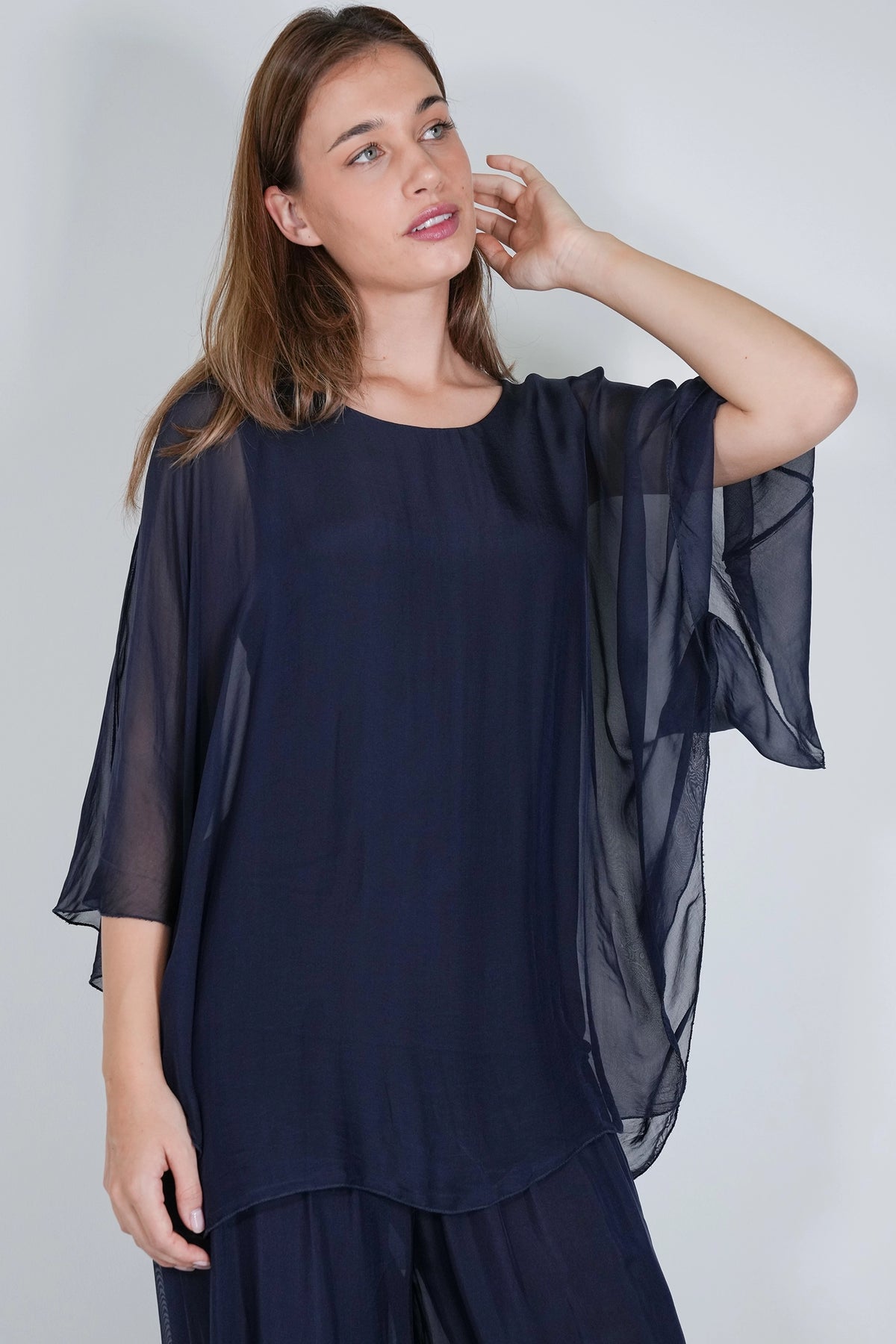 A woman wearing a navy blue sheer long-sleeve top with wide flutter sleeves and matching navy pants, photographed against a plain light gray studio background.