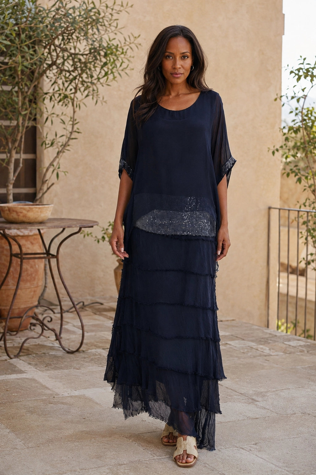 A woman modeling a dark navy blue two-piece set consisting of a short-sleeve tunic top with a sheer mesh overlay and a matching tiered maxi skirt, set against a warm beige stucco wall background.